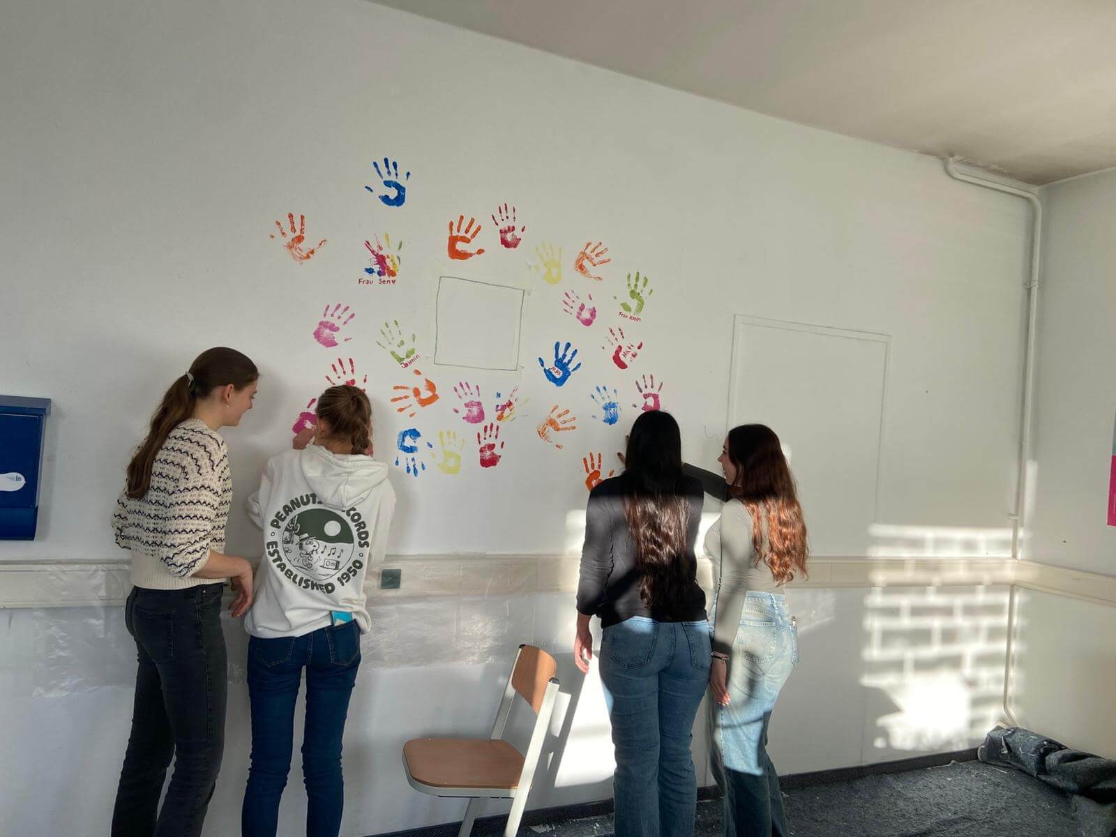 Vier junge Frauen dekorieren eine Wand mit bunten Handabdrücken in einem sonnendurchfluteten Raum.