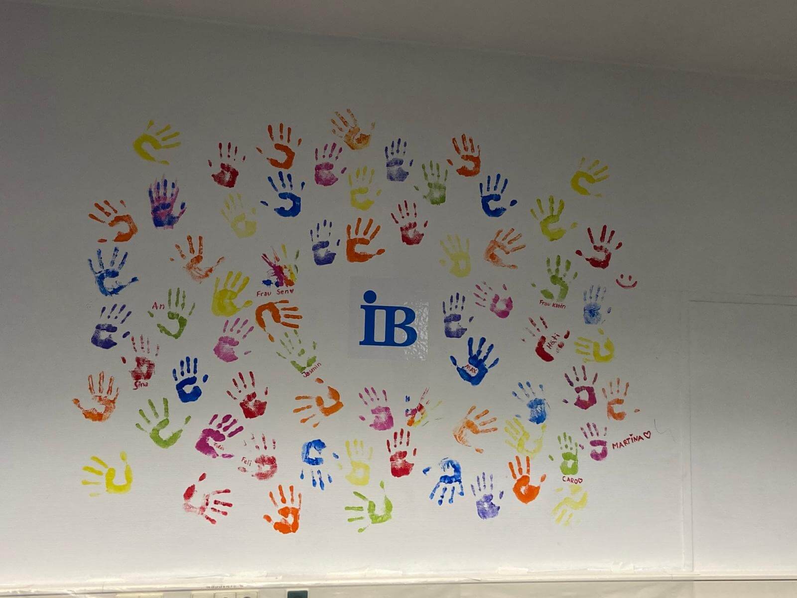 Bunte Handabdrücke in verschiedenen Farben umgeben das blaue "iB"-Logo auf einer weißen Wand.