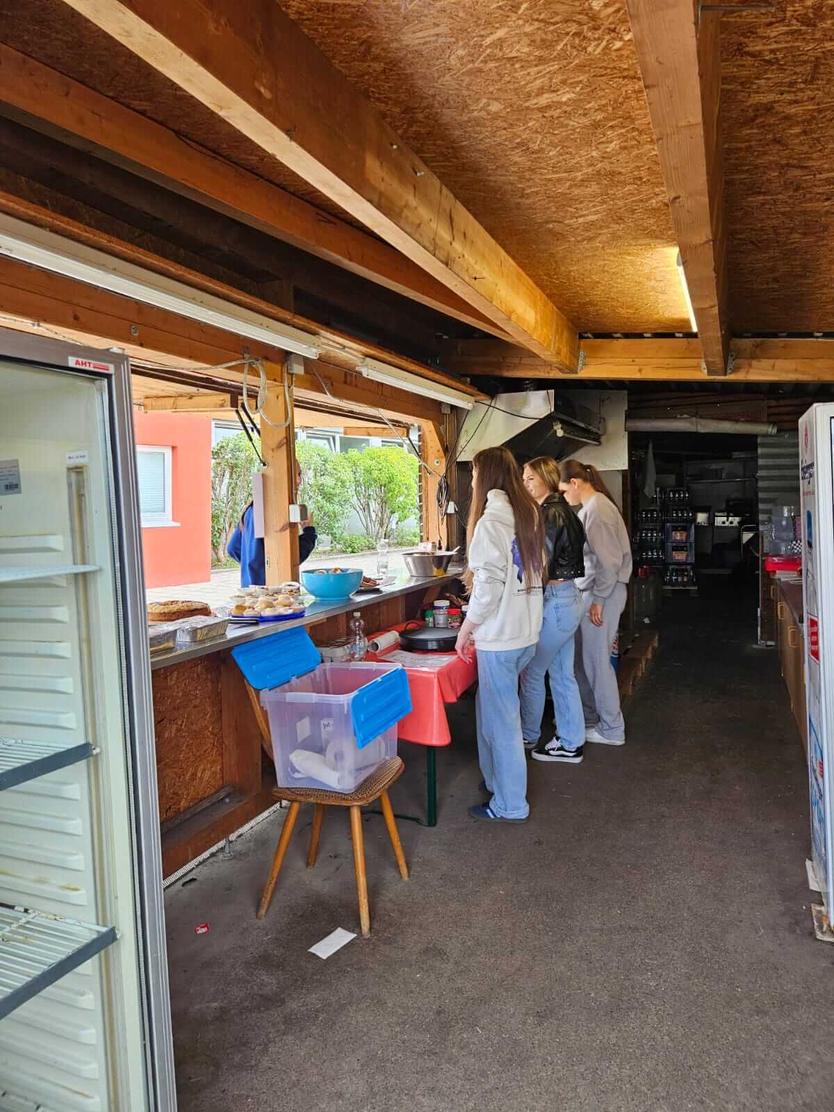 Drei Personen stehen an einem improvisierten Verkaufsstand mit Kuchen und Getränken in einem offenen Holzpavillon.