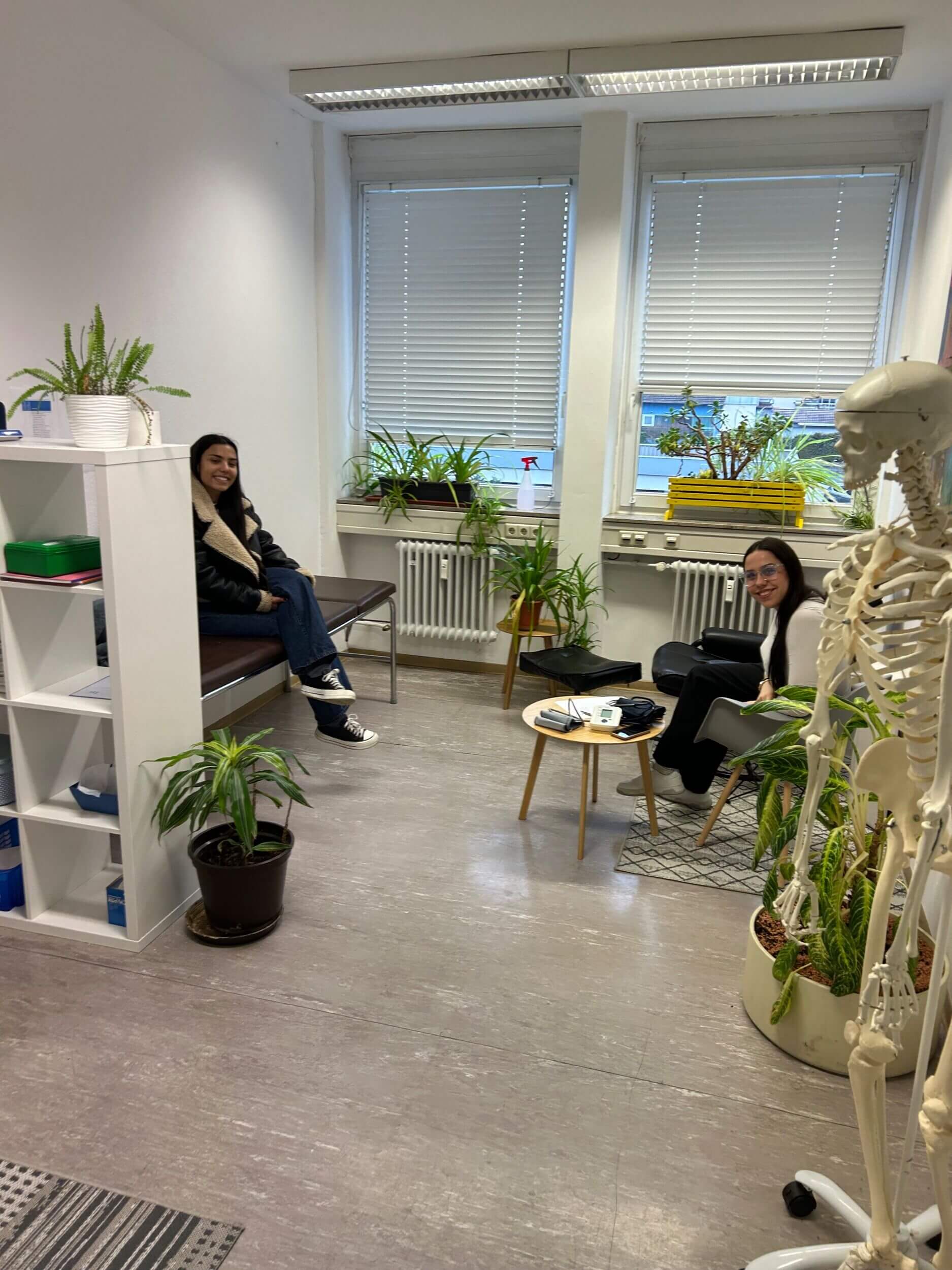 Zwei Personen sitzen in einem Wartezimmer voller Pflanzen und einem Skelett, während sie entspannt lächeln.